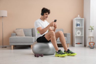 Evde modern cep telefonu ve fitness topu olan kulaklıklı genç bir adam.