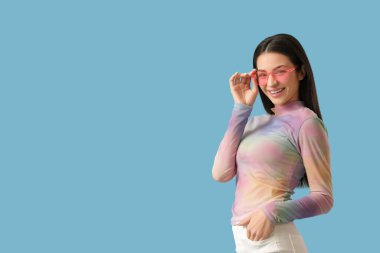 Mavi arka planda güneş gözlüklü, gülümseyen güzel genç bir kadın.