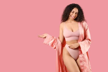 Zarif iç çamaşırı ve ipek bornoz giymiş güzel, mutlu bir Afro-Amerikalı kadın pembe arka planda bir şeyi işaret ediyor.