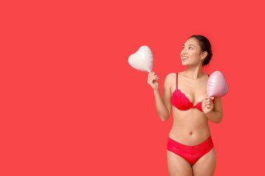 Kırmızı arka planda kalp şeklinde balonları olan seksi iç çamaşırlı güzel, mutlu bir Asyalı kadın. Sevgililer Günü kutlaması