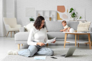 Cute little baby and young mother working with laptop while reading documents on maternity leave at home clipart