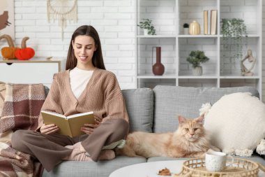Kanepede oturan genç kadın sevimli Maine Rakun kedisi ve evde kitap okuyor.
