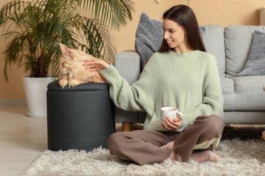 Bir fincan sıcak çay ve evde yerde oturan sevimli Maine Rakun kedisiyle genç bir kadın.