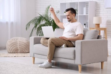 Kanepede oturan yakışıklı genç adam klima uzaktan kumandalı ve oturma odasında dizüstü bilgisayar kullanıyor.