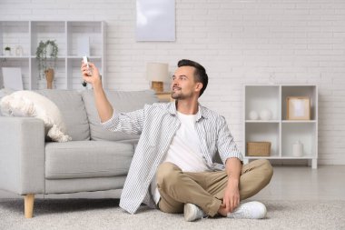 Klima kumandalı yakışıklı genç adam oturma odasında yerde oturuyor.