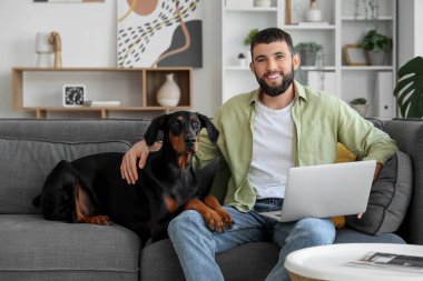 Dobermann köpeği ve dizüstü bilgisayarı olan genç adam evdeki kanepede oturuyor.