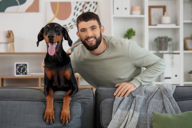 Dobermann köpekli genç adam evdeki kanepeye yaslanıyor.
