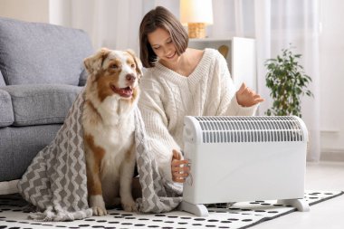 Evde ısıtıcının yanında battaniyeli genç kadın ve Avustralyalı çoban köpeği oturuyor.