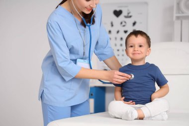 Steteskoplu kadın çocuk doktoru klinikteki kanepede küçük bir çocuğu dinliyor.