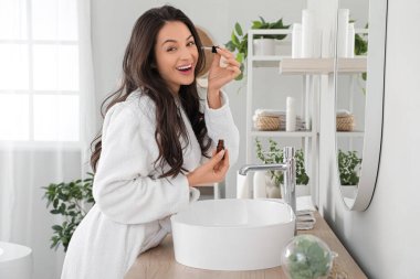 Güzel, genç, mutlu bir kadın banyoda aynanın yanında kozmetik yağı sürüyor.