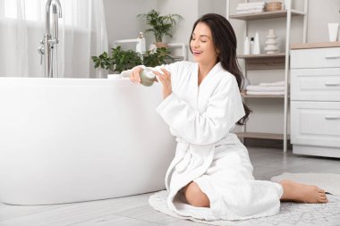 Bir kavanoz kremayla banyoda oturan güzel, genç ve mutlu bir kadın.