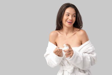 Gri arka planda pamuklu ped ve bir şişe kozmetik ürünü olan bornozlu güzel, genç bir kadın.