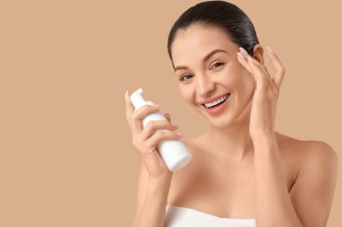 Bej arka planda bir şişe kozmetik ürünü olan güzel, genç bir kadın.