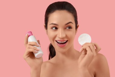 Pamuklu ped ve pembe arka planda bir şişe kozmetik ürünü olan güzel, genç bir kadın.