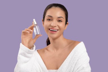 Mor arka planda bir şişe kozmetik yağıyla bornozlu güzel, genç ve mutlu bir kadın.