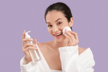 Bornozlu, güzel, genç ve mutlu bir kadın. Üzerinde bir şişe kozmetik ürünü ve mor arka planda pamuk pedi var.