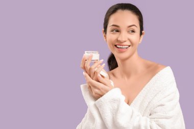 Mor arka planda krema kavanozu olan bornozlu güzel, genç ve mutlu bir kadın.