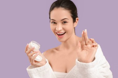 Mor arka planda krema kavanozu olan bornozlu güzel, genç ve mutlu bir kadın.