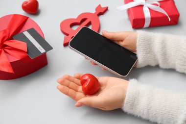 Modern cep telefonlu kadın elleri, kırmızı kalp, kredi kartı ve gri arkaplanda Sevgililer Günü için hediyeler.