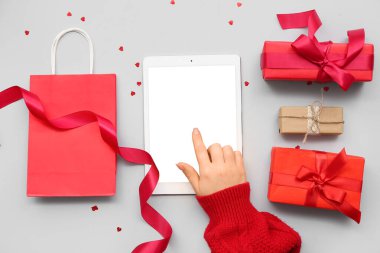 Modern tablet bilgisayarlı kadın eli, alışveriş çantası ve gri arkaplanda Sevgililer Günü için hediyeler.