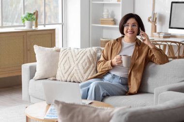 Bilgisayarlı olgun bir kadın evdeki koltukta cep telefonuyla konuşuyor.