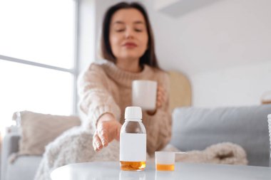 Sick young woman taking cough syrup from table at home, closeup clipart