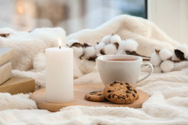 Çay fincanıyla yanan mum ve pencerenin yanındaki ekose üzerinde kurabiyeler, yakın plan.