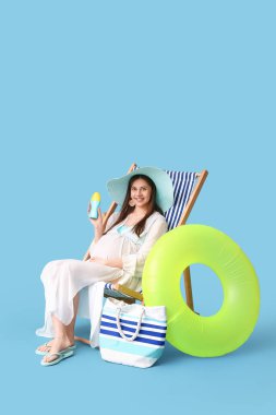 Güneş kremi şişesi ve şişirilebilir yüzükle güvertede oturan güzel hamile kadın mavi arka planda oturuyor.