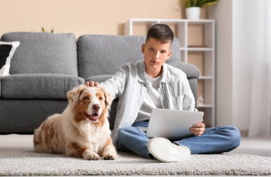 Avustralyalı çoban köpeği ve dizüstü bilgisayarı olan genç çocuk evde yerde oturuyor.
