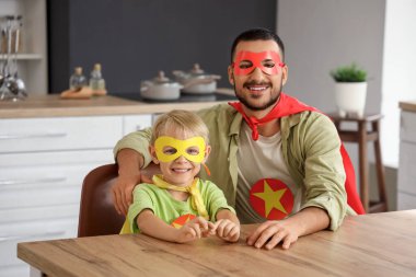 Mutlu bir baba ve süper kahraman gibi giyinmiş küçük oğlu evde mutfakta oturuyor.