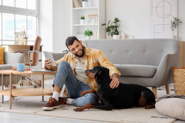 Evde tabletli genç adam ve Dobermann köpeği var.