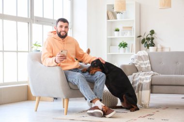 Cep telefonlu genç adam ve evdeki koltukta oturan Dobermann köpeği.