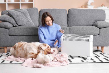 Battaniyeli genç kadın ve Avustralyalı çoban köpeği ısıtıcının yanında yerde oturuyor.