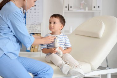 Steteskoplu kadın çocuk doktoru klinikteki kanepede küçük bir çocuğu dinliyor.
