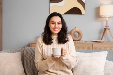 Evdeki kanepede oturan bir fincan sıcak çikolatalı güzel, genç ve mutlu kadın.