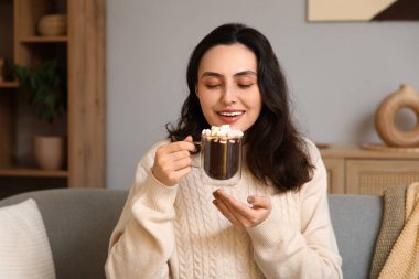 Evdeki kanepede oturan bir fincan sıcak çikolata ve marşmelovla güzel, genç ve mutlu bir kadın.