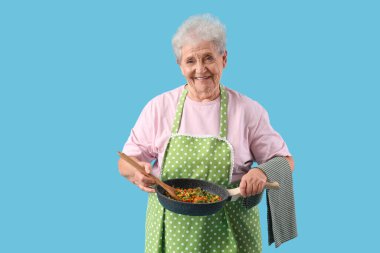 Mavi arka planda kızartma tavasında sebzeli son sınıf kadını.