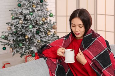 Güzel, mutlu, Asyalı bir kadın sıcak şarap ve ekoseyle evdeki Noel ağacının yanındaki kanepede oturuyor.