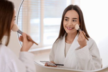 Banyoda aynanın yanında makyaj yapan genç bir kadın.