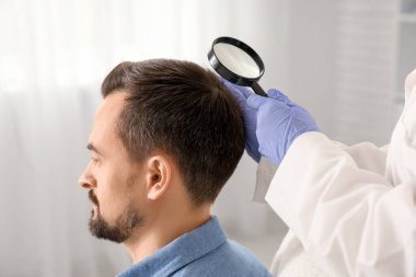 Büyüteçle trikoloğum klinikte adamın saçını inceliyor.