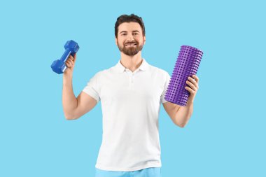 Mavi arka planda köpük silindirli ve dambılı erkek fizyoterapist