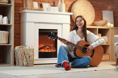 Genç bir kadın akşam şöminenin yanında gitar çalıyor.