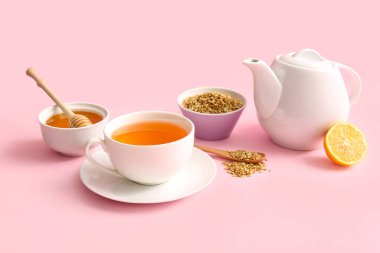 Bir fincan sıcak karabuğday çayı, limon ve bir kase bal, pembe arka planda.