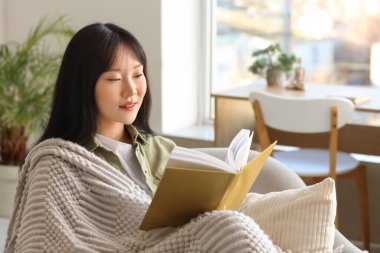 Evde gri ekose kitap okuyan genç Asyalı kadın.