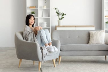 Evdeki koltukta oturan gri ekose ve kakaolu genç Asyalı kadın.