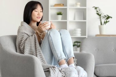 Evdeki koltukta oturan gri ekose ve kakaolu genç Asyalı kadın.