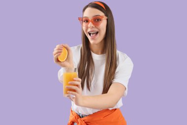 Güneş gözlüklü genç bir kadın arkaplanda portakal suyunu cama sıkıyor.