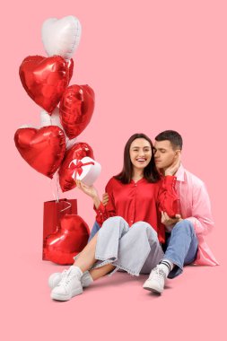 Young couple with gift and heart-shaped balloons sitting on pink background. Valentine's Day celebration