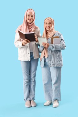 Mature Muslim women in hijabs with books and coffee on blue background clipart