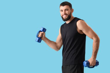 Sportif genç adam mavi arka planda dambıllarla egzersiz yapıyor.